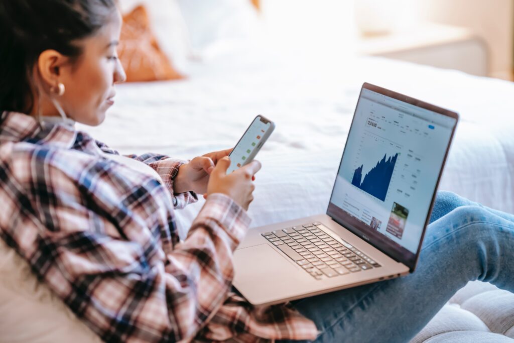 mulher latina com celular na mão e computador mostrando gráficos para representar blogpost sobre controle financeiro